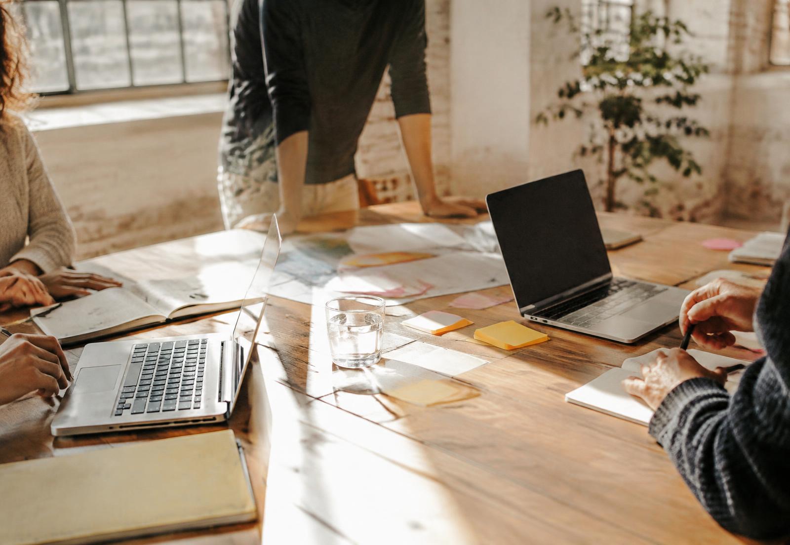A small team working together at a sunlit wooden table with laptops, notebooks and sticky notes